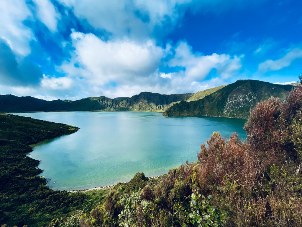 Turquoise Lagoa do Fogo surrounded by the caldera against a deep blue sky and some white clouds.