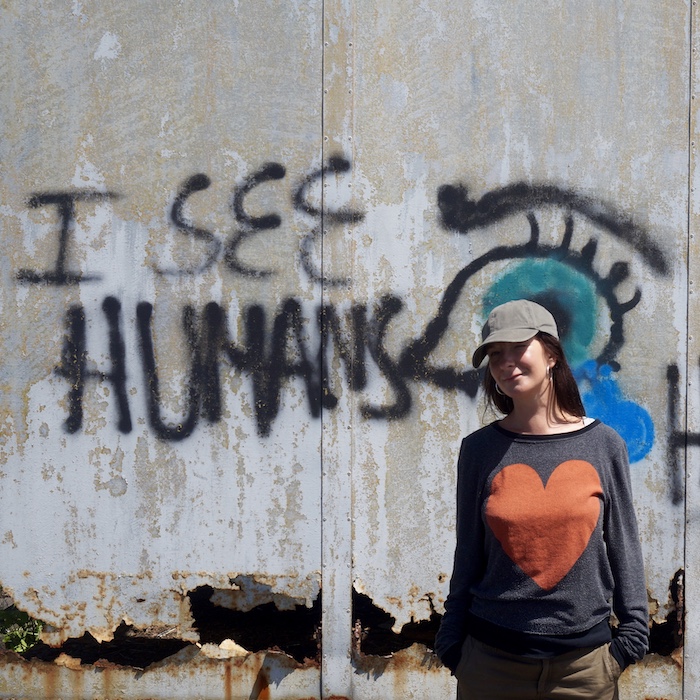 Ida Rust wearing a black sweater with a red heart, standing next to a graffiti text on a decayed wall saying I see humans